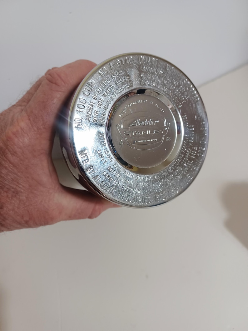 May include: A silver-colored Aladdin Stanley thermos cup. The lid is visible, with embossed text including "No. 100 Cup" and "Aladdin Stanley." The cup is held by a person.