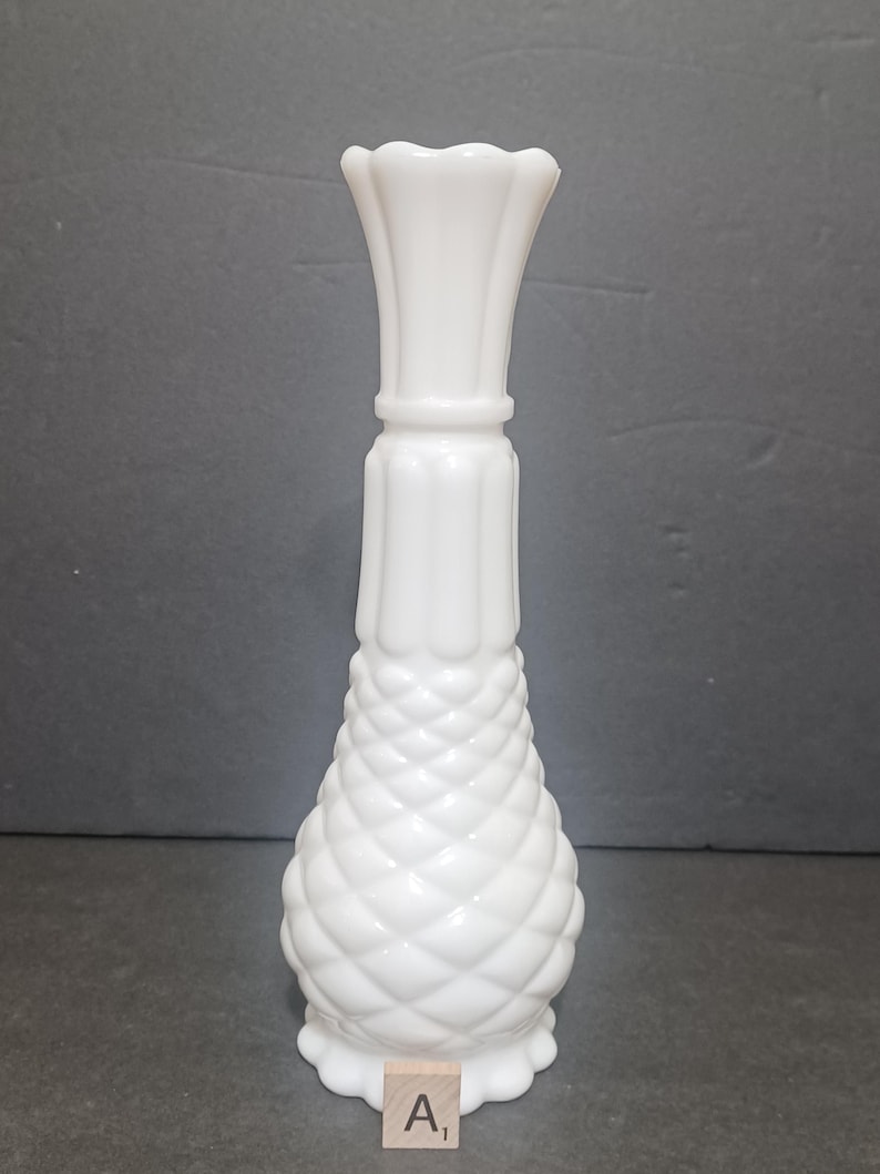 May include: A white milk glass vase featuring a textured diamond pattern on the lower bulbous section, a vertical ribbed neck, and a scalloped rim. A small wooden square tile with the letter A and the subscript 1 is placed at the base.