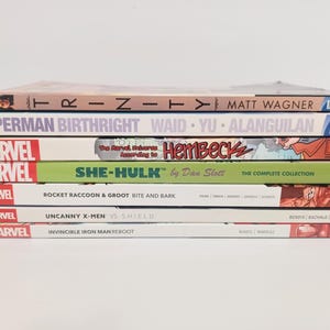 May include: A stack of comic books featuring titles like "Trinity," "Superman Birthright," and "She-Hulk." The books have colorful covers with various Marvel and DC Comics logos. The spines display author names and series titles.