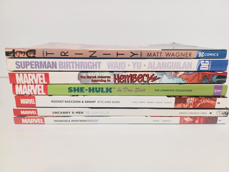 May include: A stack of comic books featuring titles like "Trinity," "Superman Birthright," and "She-Hulk." The books have colorful covers with various Marvel and DC Comics logos. The spines display author names and series titles.
