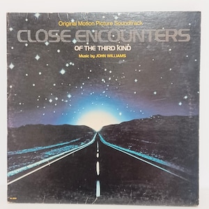 May include: Vintage vinyl record album cover for "Close Encounters of the Third Kind" soundtrack. The cover features a dark blue night sky with a road leading towards a bright light. Text includes the title and "Music by John Williams."