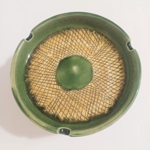 May include: A round ceramic ashtray with a green rim and a textured yellow and black center. The center has a raised green circle in the middle. The ashtray has three small notches for cigarettes.