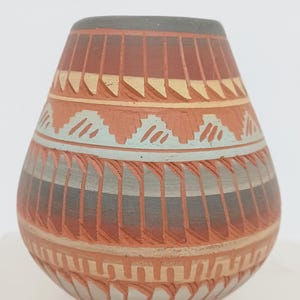 May include: A rounded ceramic vase featuring a terracotta base color with horizontal bands of geometric patterns in shades of dark gray, cream, and light blue. The surface displays carved vertical lines and stepped motifs encircling the body.