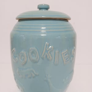 May include: A light blue ceramic cookie jar with a lid. The word "COOKIES" is embossed on the front in a lighter color. The jar has embossed animal designs around the base.