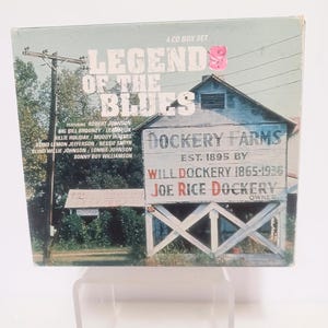 May include: A CD box set titled "Legends of the Blues" featuring Robert Johnson, Billie Holiday, and others. The cover shows a rural scene with a building that reads "Dockery Farms" and the text "Est. 1895".