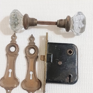 May include: A set of door hardware including two clear glass knobs connected by a metal spindle, two elongated brass keyhole escutcheon plates with decorative finials, and a rectangular black metal mortise lock box with a brass faceplate.