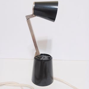 May include: A black desk lamp with a silver adjustable arm and a conical shade. The lamp has a black base and a white power cord. The lamp is designed for desk use and provides focused lighting.