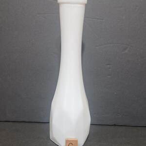 May include: A tall, white milk glass bud vase with a slender neck and a flared, faceted base. The rim features a delicate scalloped edge. A small, square wooden tile with the letter C and the number 3 is positioned at the base of the vase.