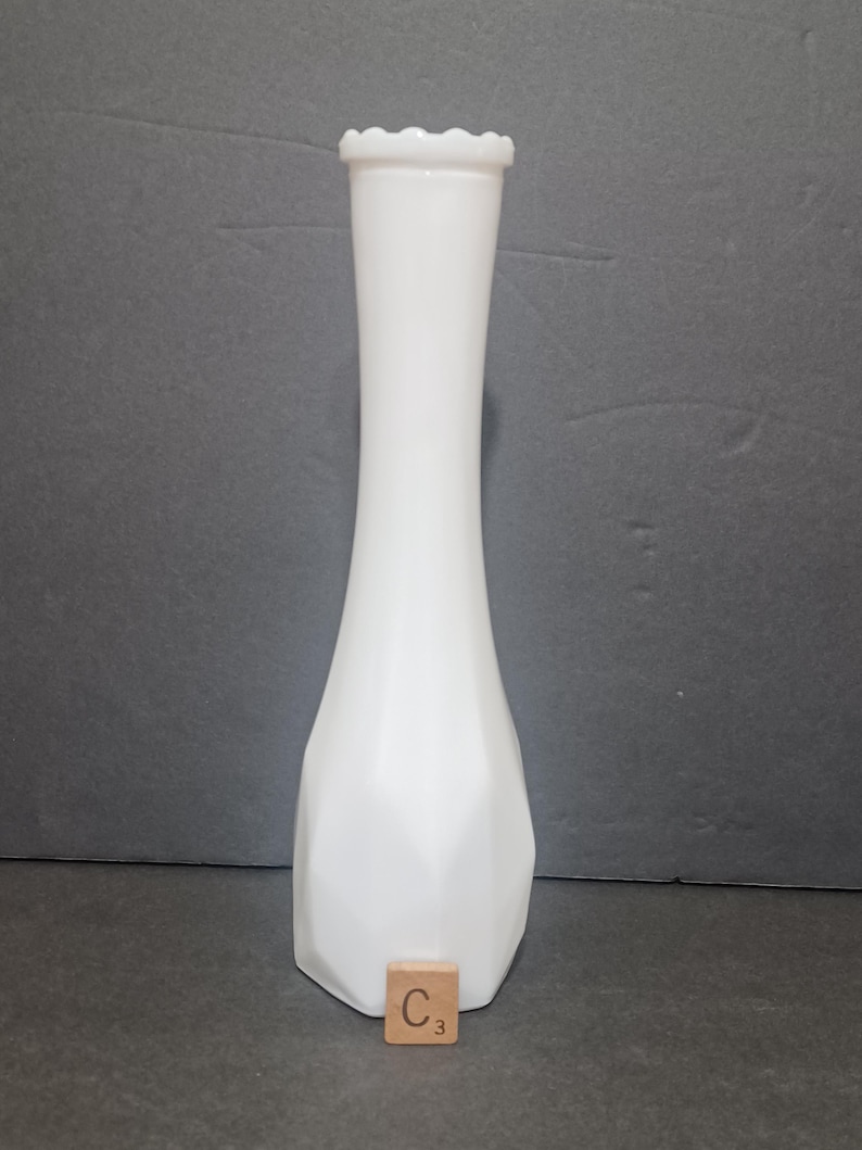 May include: A tall, white milk glass bud vase with a slender neck and a flared, faceted base. The rim features a delicate scalloped edge. A small, square wooden tile with the letter C and the number 3 is positioned at the base of the vase.