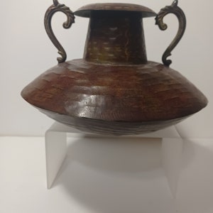May include: A brown metal vase with two ornate handles and a hammered texture. The vase is sitting on a white stand.