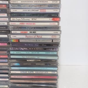 May include: A vertical stack of music CDs in clear plastic jewel cases. The spines display various artist names and album titles including Foreigner, Glenn Frey, Jackson Browne, John Mellencamp, Journey, Kid Rock, Loverboy, Neil Young, R.E.M., and Yes.