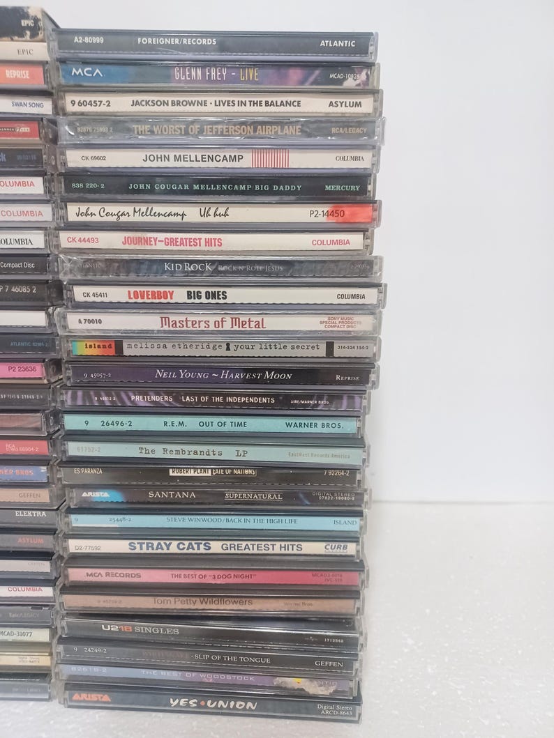 May include: A vertical stack of music CDs in clear plastic jewel cases. The spines display various artist names and album titles including Foreigner, Glenn Frey, Jackson Browne, John Mellencamp, Journey, Kid Rock, Loverboy, Neil Young, R.E.M., and Yes.