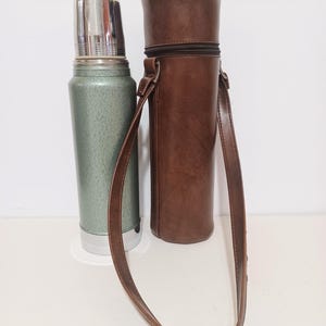 May include: A vintage green and silver thermos with a stainless steel lid, next to a brown leather carrying case with a shoulder strap. The thermos is cylindrical, and the case has a zipper closure.