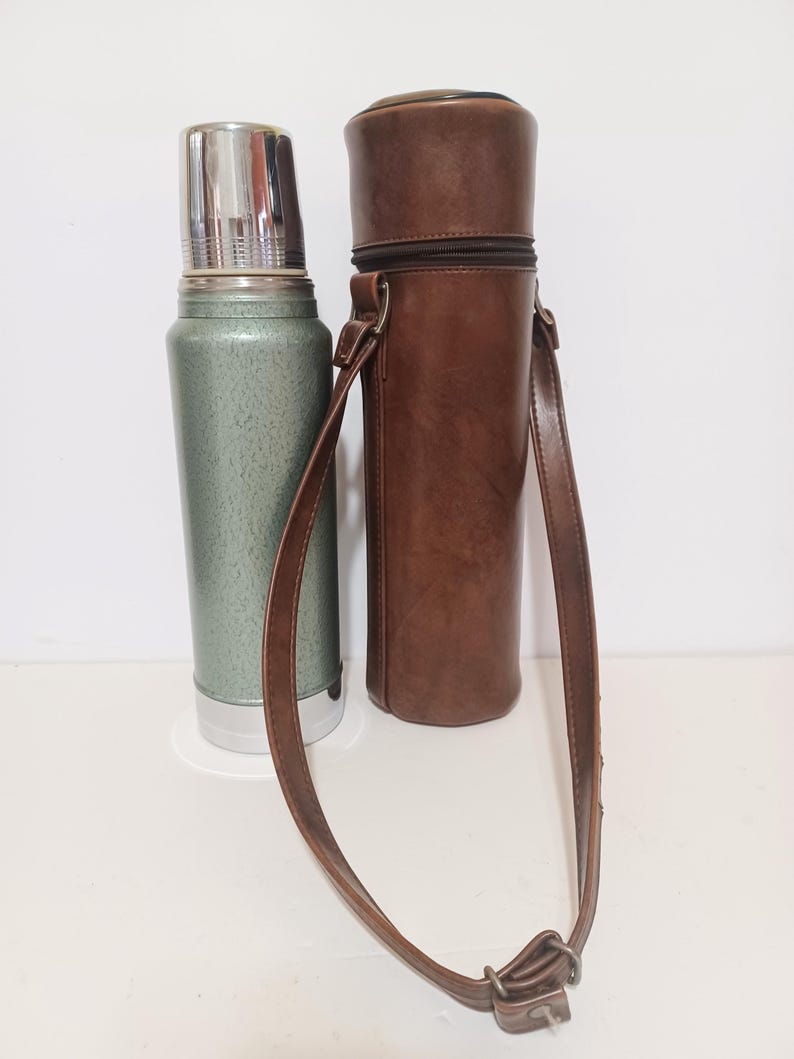May include: A vintage green and silver thermos with a stainless steel lid, next to a brown leather carrying case with a shoulder strap. The thermos is cylindrical, and the case has a zipper closure.