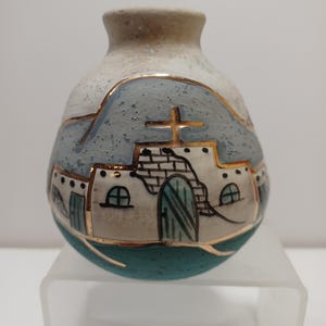 May include: A small, round ceramic vase with a blue, white, and gold painted design of a pueblo-style building with a cross on top. The vase has a textured surface and a teal green base.