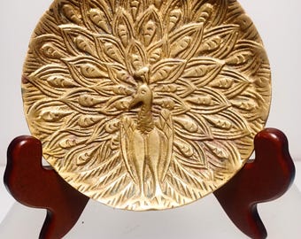 May include: A gold-colored brass dish with an embossed peacock design. The peacock is in the center of the dish with its tail feathers spread out. The dish is on a wooden stand.