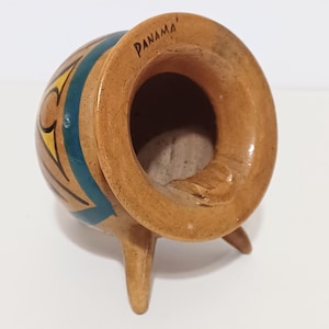May include: A small, handcrafted ceramic pot with three legs. The pot is tan with blue and yellow geometric designs and the word "PANAMA" is written on the rim. The pot has a wide opening and a rough interior.