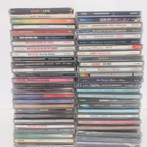 May include: Two vertical stacks of music CDs in plastic jewel cases. The spines display various artist names and album titles including AC/DC, Bob Dylan, Bruce Hornsby, The Doors, Foreigner, Jackson Browne, John Mellencamp, and Yes, among others.