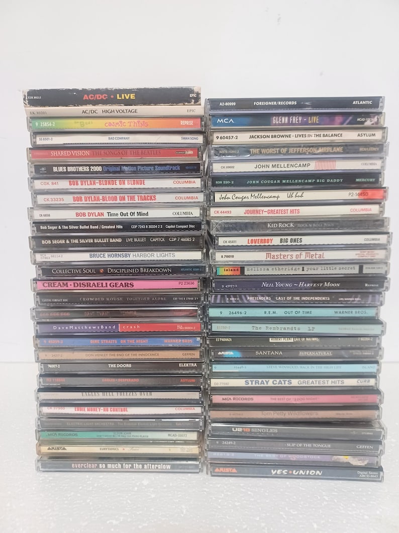 May include: Two vertical stacks of music CDs in plastic jewel cases. The spines display various artist names and album titles including AC/DC, Bob Dylan, Bruce Hornsby, The Doors, Foreigner, Jackson Browne, John Mellencamp, and Yes, among others.