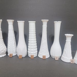 May include: A collection of seven white milk glass bud vases of varying heights and shapes, each featuring distinct embossed patterns or smooth surfaces. Each vase is labeled with a small wooden letter tile from A to G at its base.