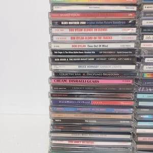 May include: A vertical stack of numerous music compact discs in plastic jewel cases. The spines are visible, displaying various artist names and album titles including AC/DC, Bob Dylan, Bob Seger, Bruce Hornsby, Cream, and Everclear in various colors.