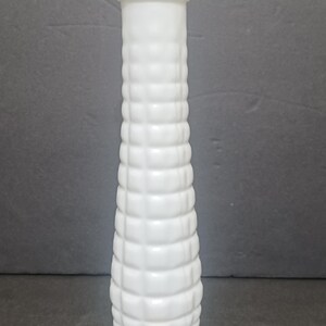 May include: A tall, cylindrical white vase with a textured, grid-like pattern of raised squares covering its exterior surface. The vase features a smooth, flat rim at the top and a small, square wooden tile labeled D2 placed at its base.
