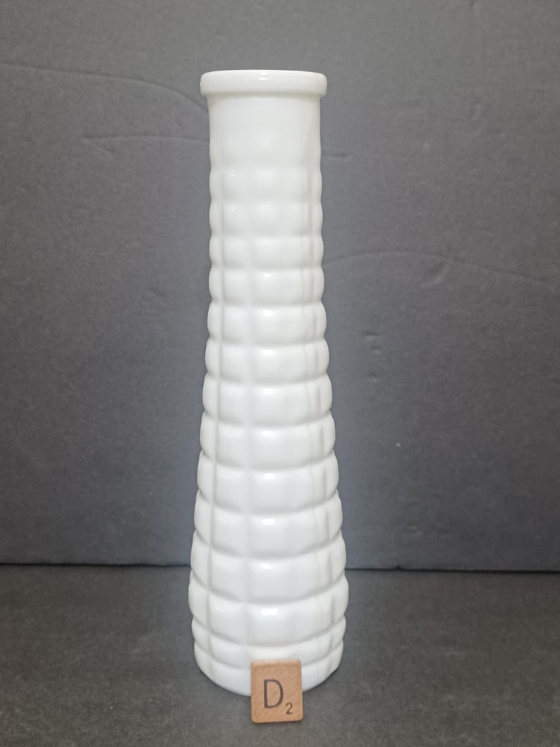 May include: A tall, cylindrical white vase with a textured, grid-like pattern of raised squares covering its exterior surface. The vase features a smooth, flat rim at the top and a small, square wooden tile labeled D2 placed at its base.