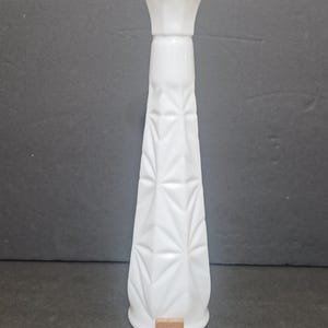 May include: A tall, white milk glass bud vase featuring a tapered, geometric starburst pattern along its body and a flared, octagonal rim. A small wooden square tile with the letter B and the number 3 is positioned at the base of the vase.