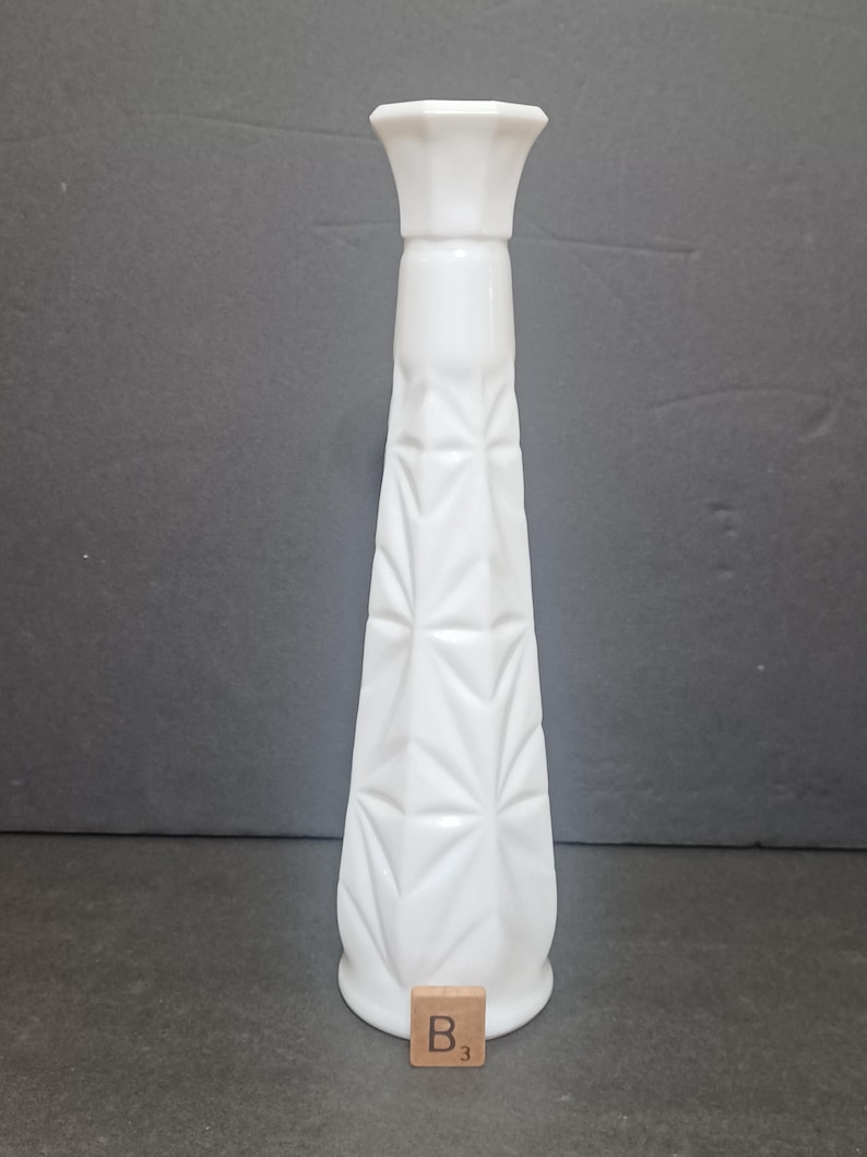 May include: A tall, white milk glass bud vase featuring a tapered, geometric starburst pattern along its body and a flared, octagonal rim. A small wooden square tile with the letter B and the number 3 is positioned at the base of the vase.