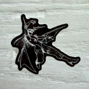 May include: A die-cut sticker featuring a black silhouette of a winged figure with long hair and pointed ears. The design is rendered in a white line art style, depicting the character in a dynamic, outstretched pose with large, sharp wings.