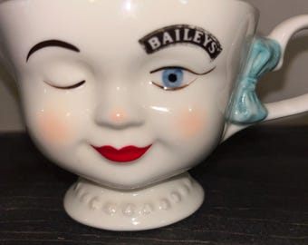 Puede incluir: Taza de cerámica blanca con forma de cara con un ojo azul, guiñando un ojo y labios rojos. La taza tiene un lazo azul en el asa y la palabra "BAILEYS" impresa en la frente.