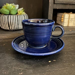 May include: A blue ceramic teacup and saucer set. The cup has a rounded shape and a handle. The saucer has a scalloped edge.