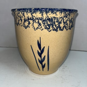 Op de afbeelding: Een beige keramische pot met een blauw gespikkelde rand en een blauw tarwe-aarontwerp. De pot heeft een afgeronde vorm en een licht uitlopende bovenkant. Het ontwerp is eenvoudig en rustiek.