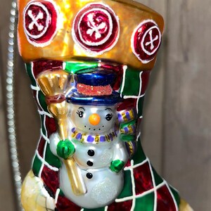 May include: A glass Christmas ornament shaped like a stocking with a snowman inside. The stocking is red, green, and gold with a plaid pattern. The snowman is wearing a blue hat and a purple scarf.