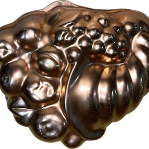 May include: A vintage copper-colored metal mold for baking a fruit-shaped cake. The mold is shaped like a cornucopia with grapes, apples, and pears.