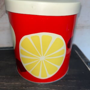 Puede incluir: Un recipiente cilíndrico rojo y blanco con un diseño de limón amarillo. El recipiente tiene una tapa blanca.