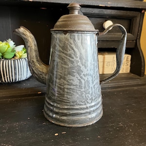 Peut inclure: Une théière en émail vintage de couleur gris et blanc moucheté avec un couvercle rouillé et une poignée noire. La théière est posée sur une surface noire.