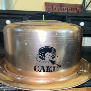 May include: A vintage silver cake dome with a black and white graphic of a cake and the word "CAKE" on the front.