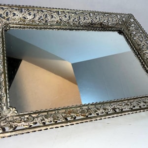 May include: An antique rectangular vanity mirror with an ornate, filigree-style metal frame. The frame is a mix of silver and gold tones, with intricate floral and scrollwork designs. The mirror reflects the surrounding environment.