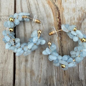 May include: A pair of hoop earrings featuring light blue tumbled stone chips and small gold-toned metal beads wired onto a circular frame. The earrings include clear plastic earring backs and exhibit a textured, multi-faceted appearance.