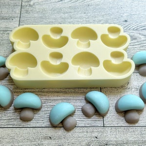 Puede incluir: Un molde de silicona para hacer caramelos o jabones con forma de hongo. El molde es de color amarillo claro y tiene ocho cavidades con forma de hongo. También hay ocho caramelos o jabones con forma de hongo azul y gris junto al molde.