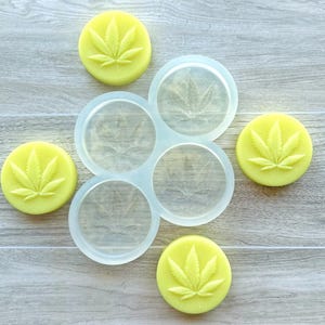 Puede incluir: Un molde de silicona transparente con cuatro impresiones circulares y tres objetos redondos amarillos. Cada objeto redondo y las impresiones del molde presentan un diseño de hoja de cannabis. Los objetos están sobre una superficie de vetas de madera clara.