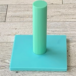 May include: A light blue, square-shaped base with a light blue cylinder standing upright in the center.