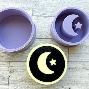 May include: A purple silicone mold for making a round soap with a crescent moon and star design. The mold is shown with a finished soap bar.