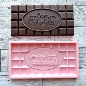 May include: A pink silicone chocolate mold with the words "Love Chocolate" in a cursive font. The mold is shaped like a chocolate bar with squares and a raised oval in the center.