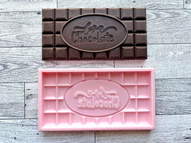 May include: A pink silicone chocolate mold with the words "Love Chocolate" in a cursive font. The mold is shaped like a chocolate bar with squares and a raised oval in the center.