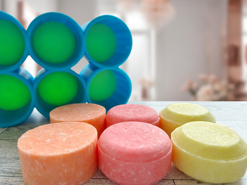 Pu&ograve; includere: Una collezione di shampoo solidi colorati, rotondi e lisci, in arancione, rosa e giallo, accompagnati da un portasapone blu e verde. Il portasapone presenta un design geometrico.