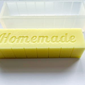 May include: A yellow rectangular soap bar with the word "Homemade" embossed on top. The soap is segmented into vertical sections. A clear, rectangular mould is in the background.