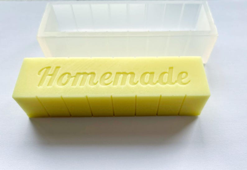 May include: A yellow rectangular soap bar with the word "Homemade" embossed on top. The soap is segmented into vertical sections. A clear, rectangular mould is in the background.