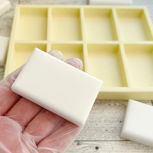 May include: A white rectangular silicone mold with six individual compartments for making soap bars. The mold is on a wooden surface, and a white soap bar is being held in a gloved hand.